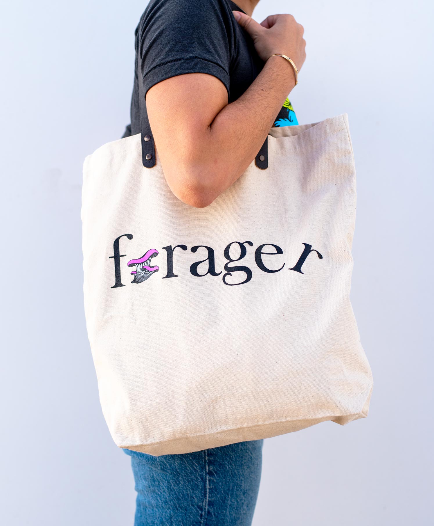 Woman holding a canvas tote bag with forager printed on the side with a mushroom for the o.